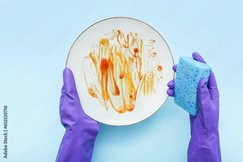 Female hands in rubber gloves washing dirty plate with sponge on blue background