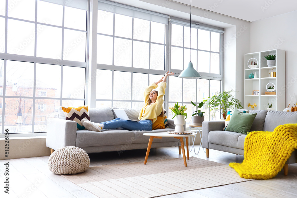 Beautiful young woman lying in cozy sofa with laptop near big window