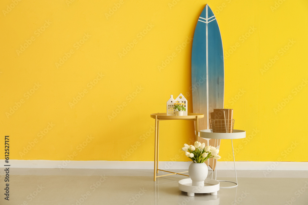 Tables with flowers, books and surfboard near color wall in room