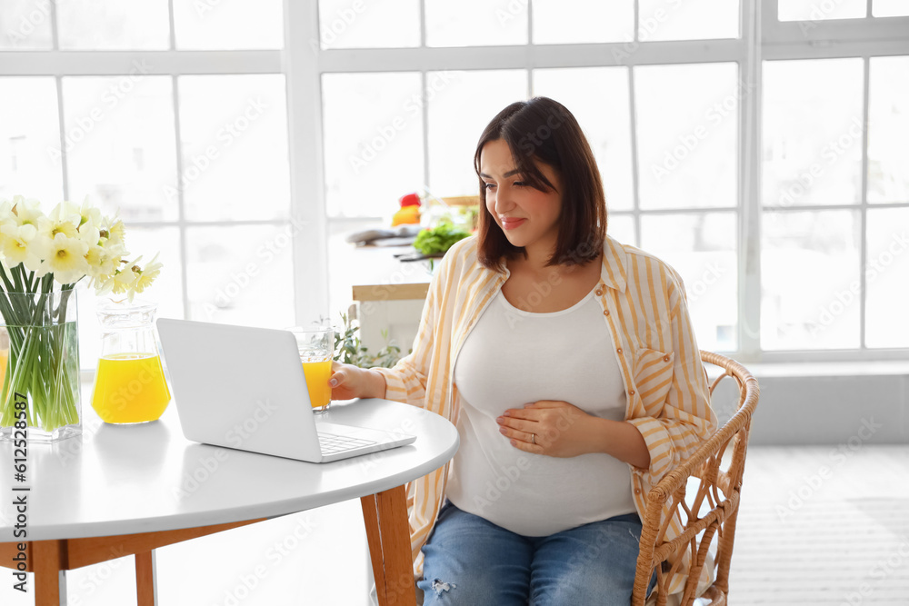 Young pregnant woman with glass of juice using laptop at table in kitchen