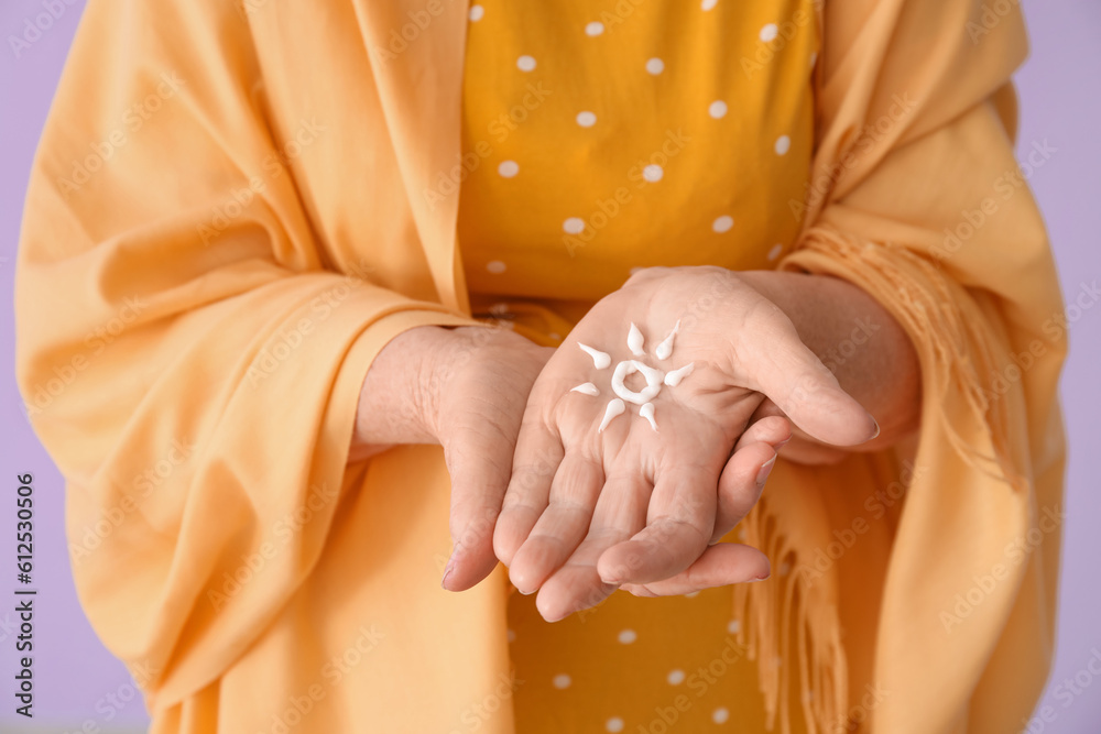 Senior woman with sun made of sunscreen cream on lilac background, closeup