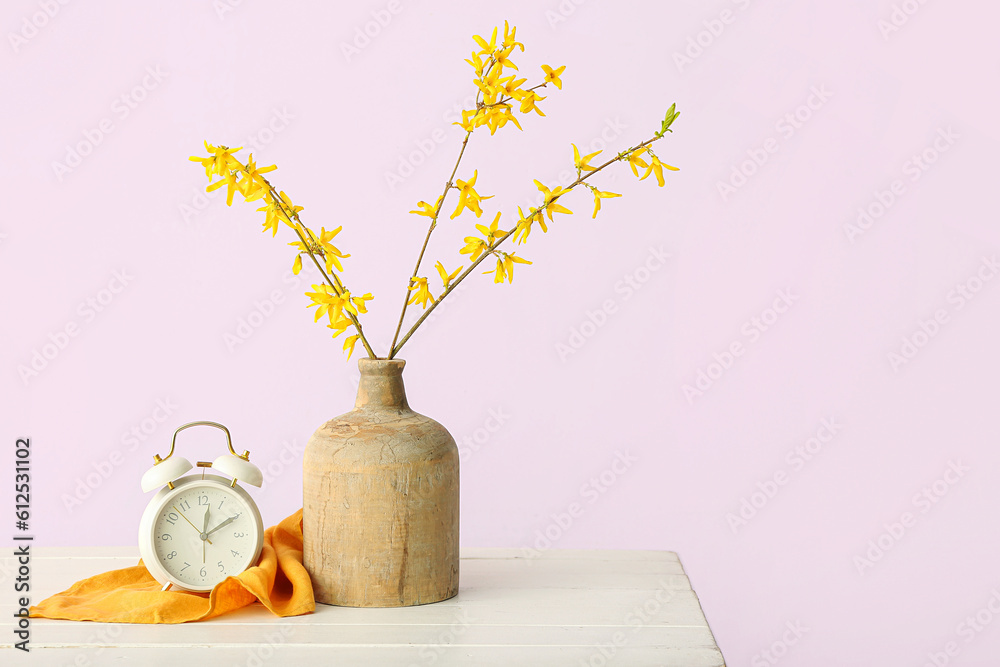 Vase with blooming tree branches and alarm clock on white table against pink background