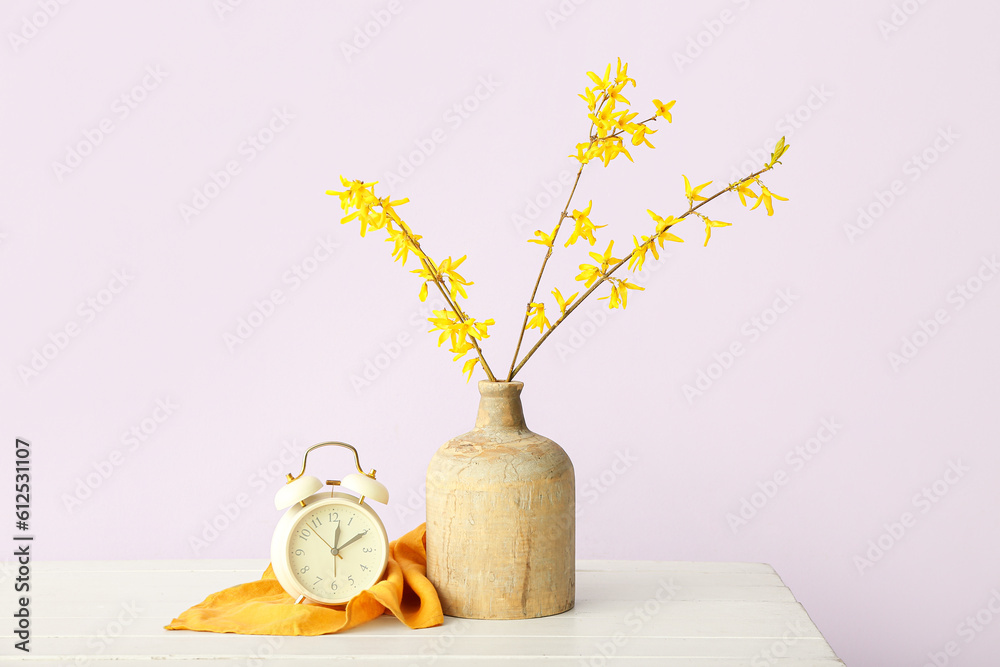 Vase with blooming tree branches and alarm clock on white table against pink background