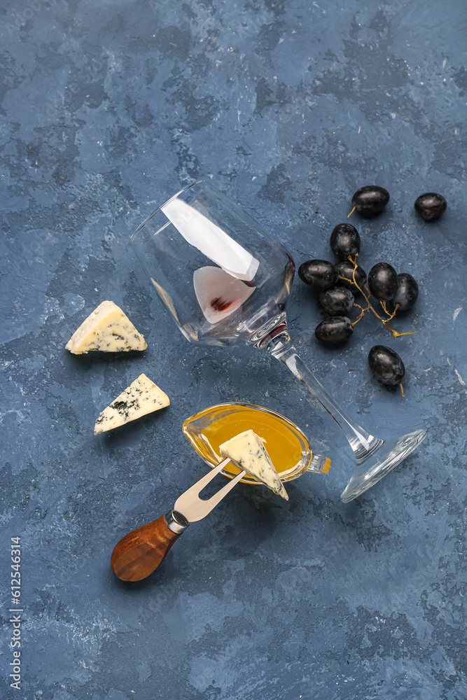 Wine glass, pieces of tasty cheese, honey and grapes on blue background