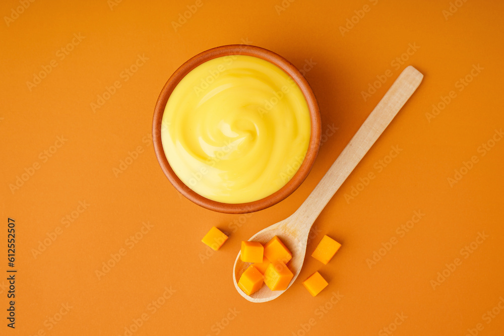 Bowl of tasty cheddar sauce and spoon with cheese cubes on orange background