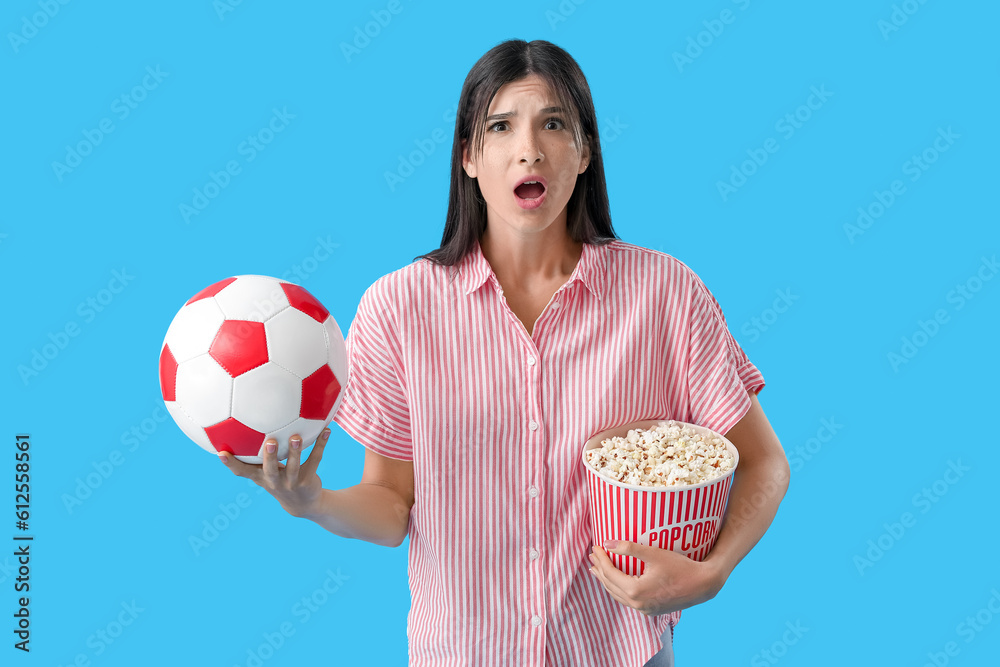 Shocked female soccer fan with popcorn and ball on blue background