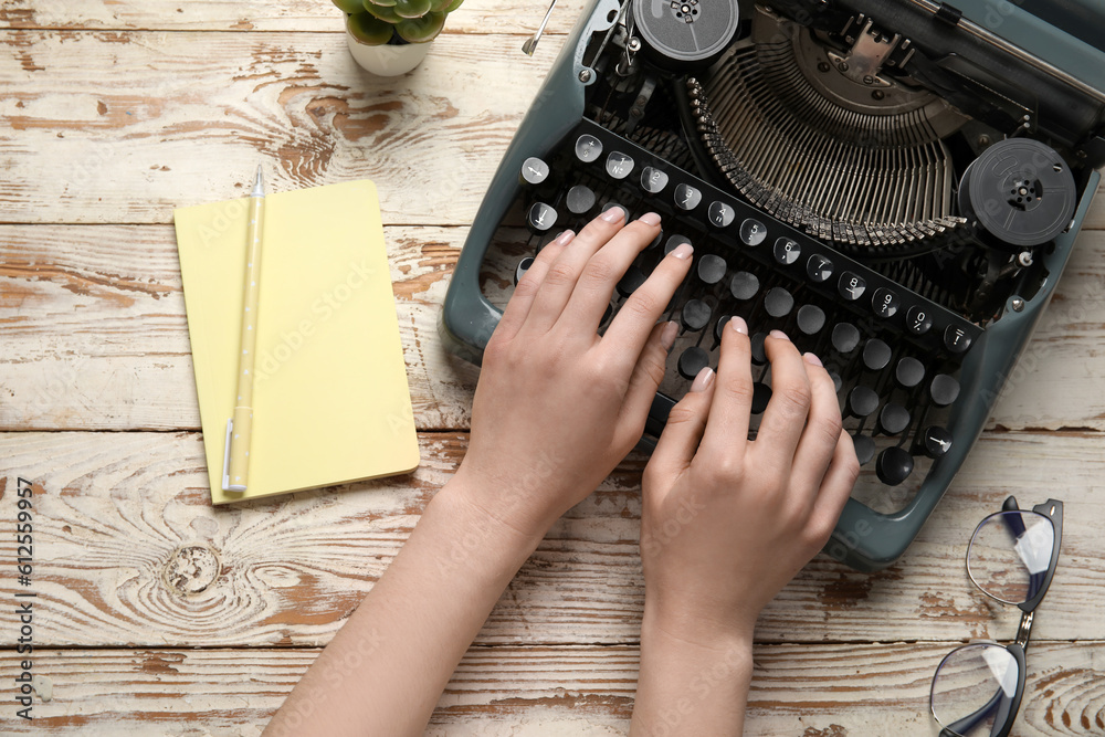 Woman typing on typewriter with eyeglasses, notebook and houseplant on wooden background