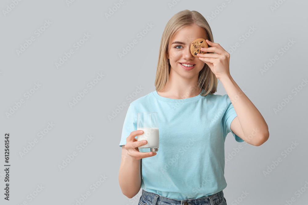 Beautiful young woman with glass of milk and cookie on grey background