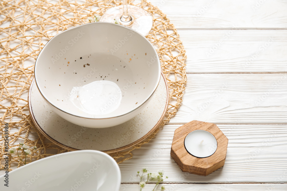 Wicker mat with plates, cutlery and candle on white wooden background