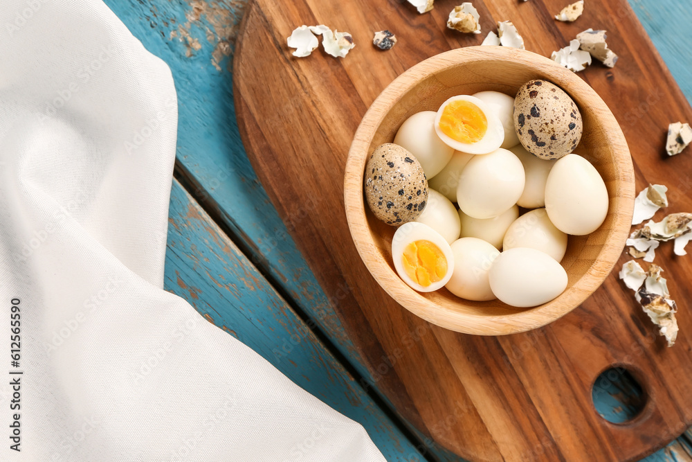 Board with bowl of boiled quail eggs and shells on blue wooden background