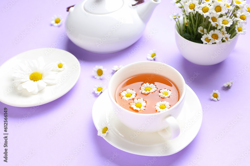 Teapot with cup of natural chamomile tea and flowers on lilac background