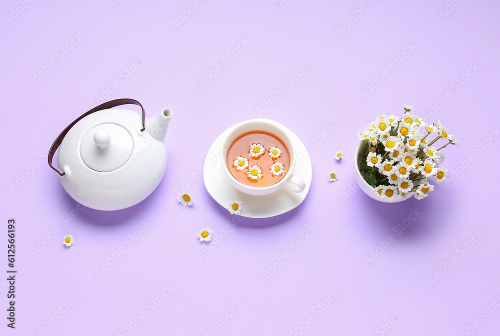 Teapot with cup of natural chamomile tea and flowers on lilac background