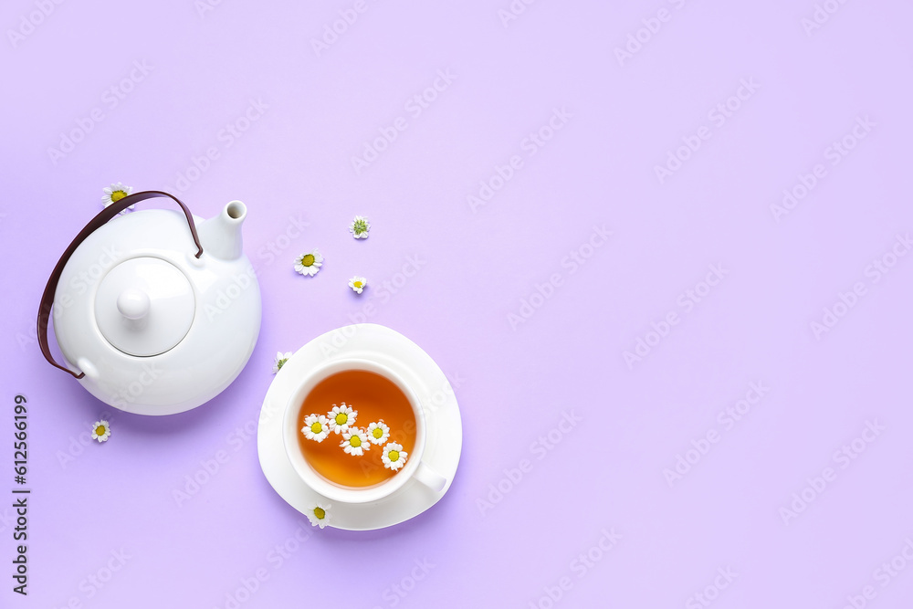 Teapot with cup of natural chamomile tea and flowers on lilac background