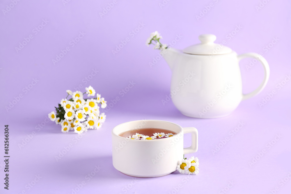 Teapot with cup of natural chamomile tea and flowers on lilac background