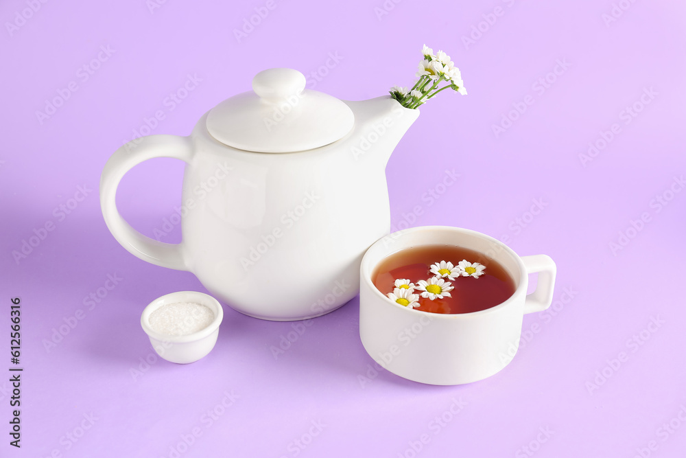 Teapot with cup of natural chamomile tea, sugar and flowers on lilac background