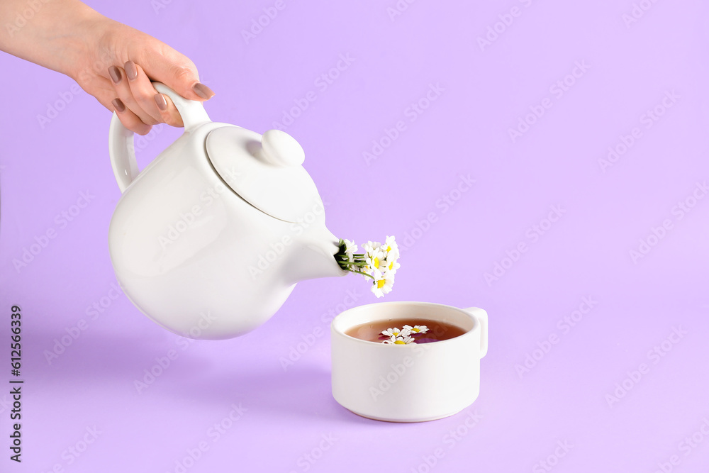 Female hand pouring natural chamomile tea from teapot into cup on lilac background