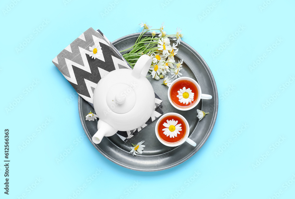 Tray with teapot, cup of natural chamomile tea and flowers on blue background