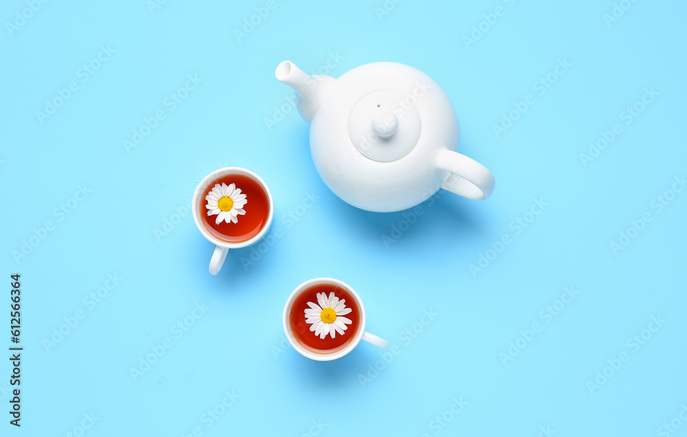 Teapot with cups of natural chamomile tea and flowers on blue background