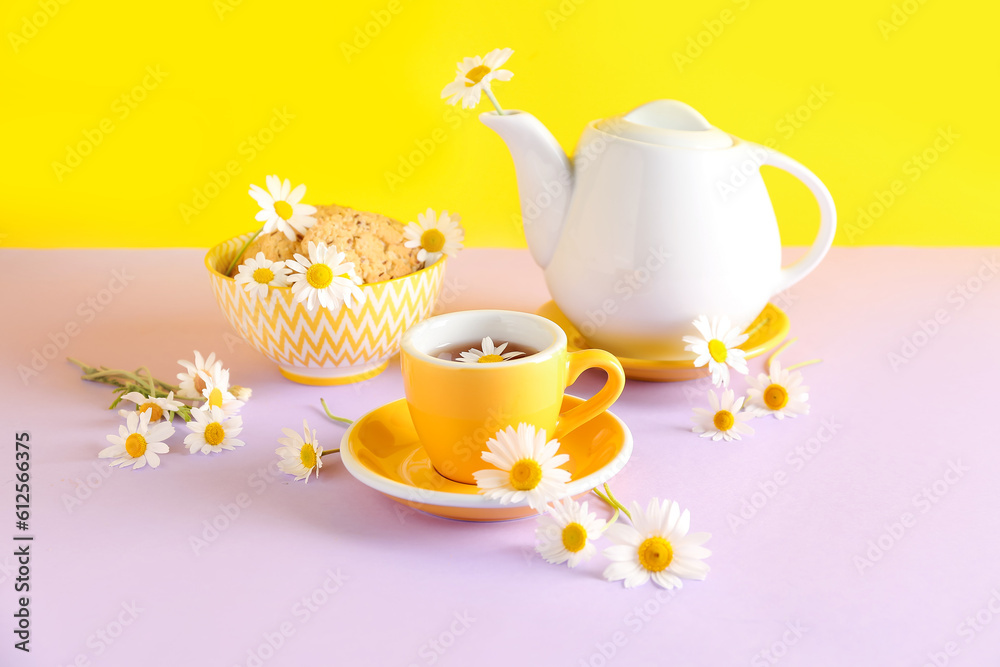 Teapot with cup of natural chamomile tea, cookies and flowers on pink table near yellow wall