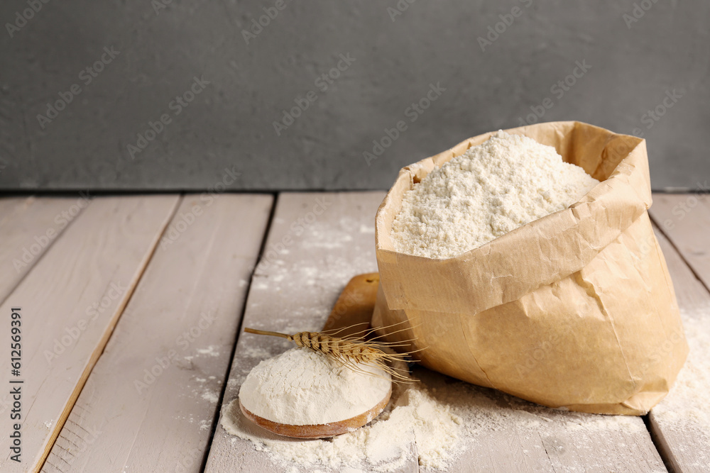 Paper bag with flour, wooden spoon and wheat ear on grey wooden table near wall