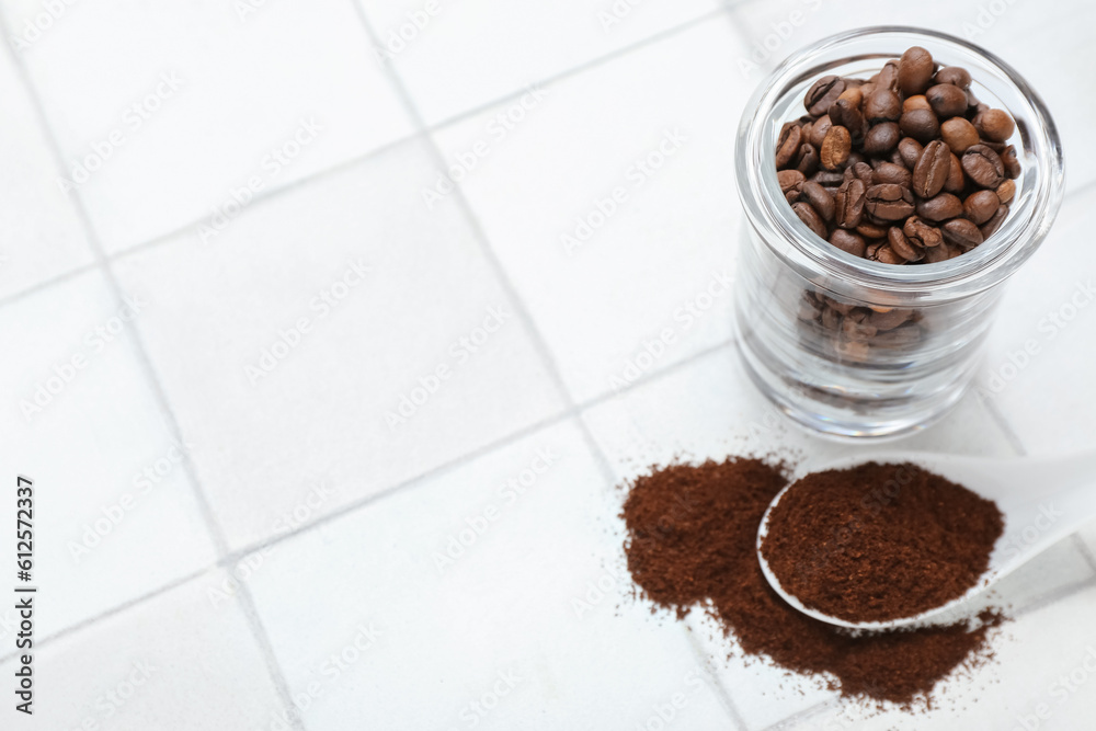 Jar with beans and spoon of coffee powder on white tile background