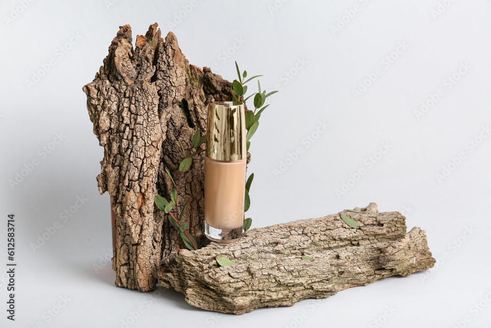 Bottle of makeup foundation, tree bark and eucalyptus branches on light background