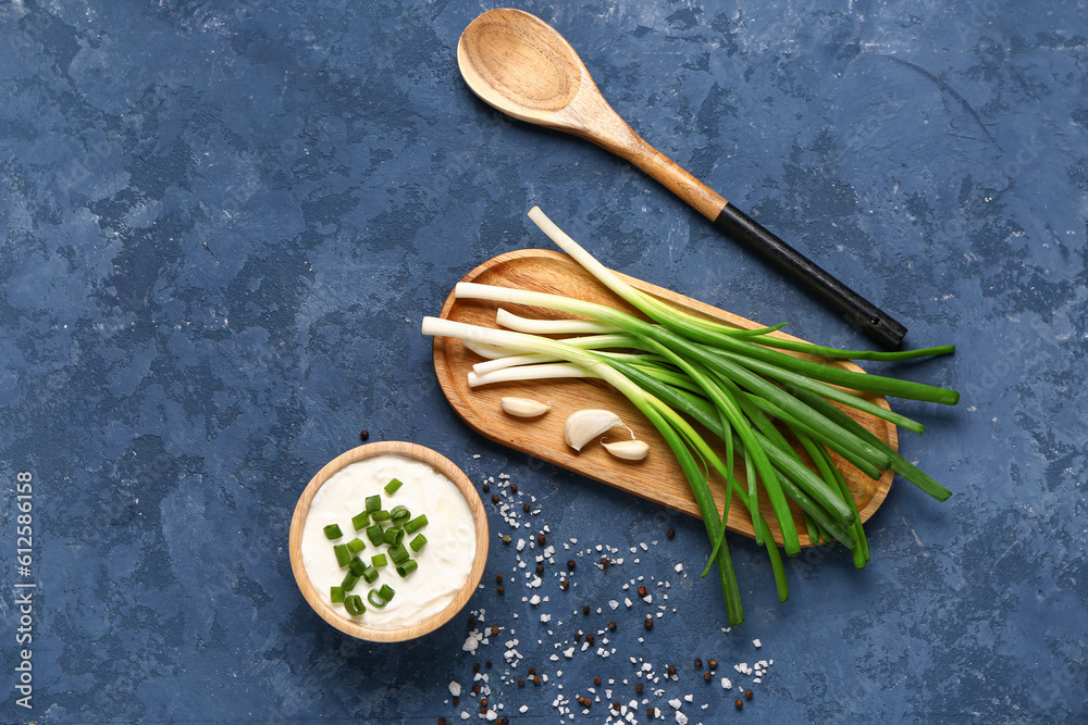 Bowl of tasty sour cream and board with green onion on blue background