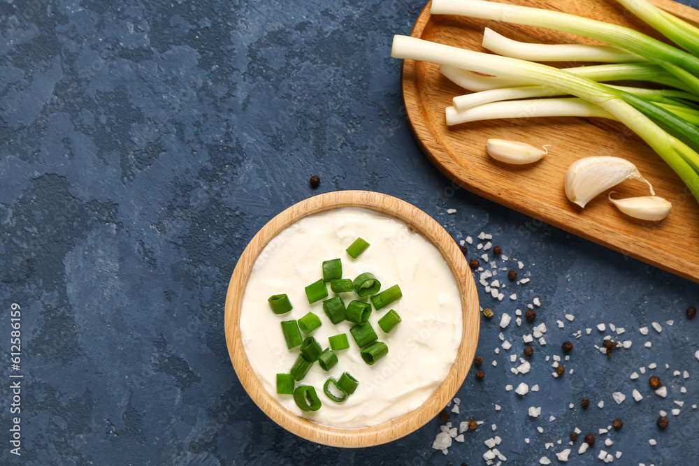 Bowl of tasty sour cream and board with green onion on blue background