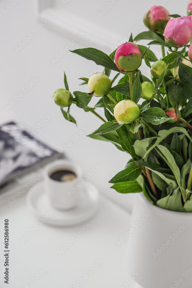 Vase with unopened peonies, cup of coffee and magazines on dresser near white wall