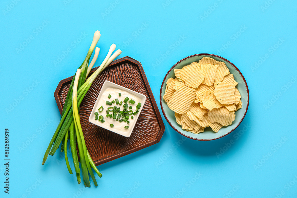 Bowl of tasty sour cream with sliced green onion and potato chips on blue background