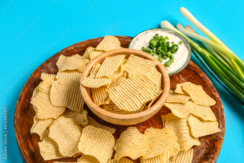 Bowl of tasty sour cream with sliced green onion and potato chips on blue background