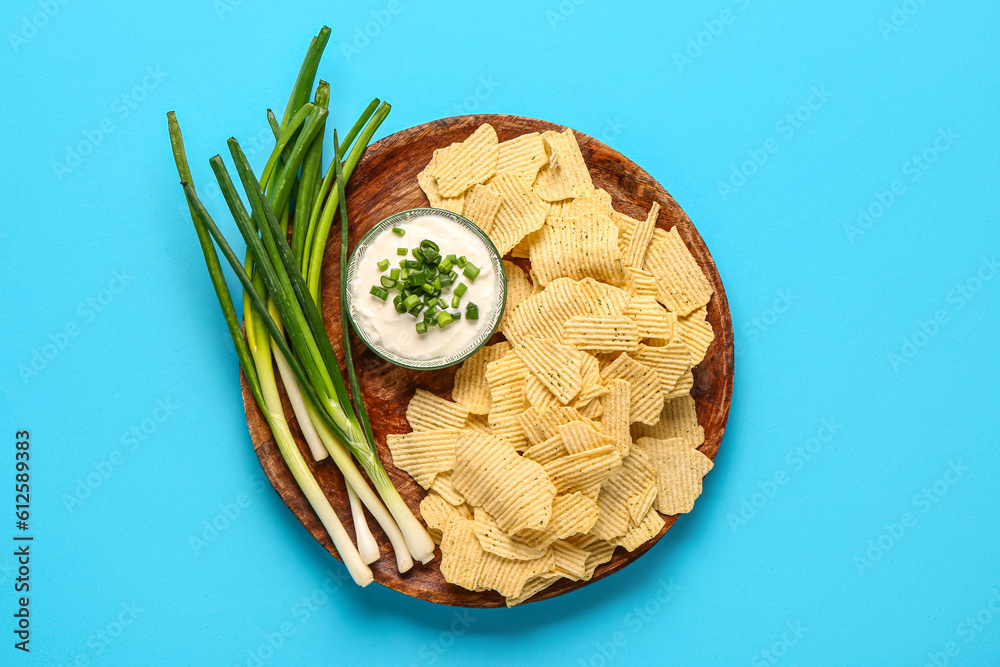 Bowl of tasty sour cream with sliced green onion and potato chips on blue background