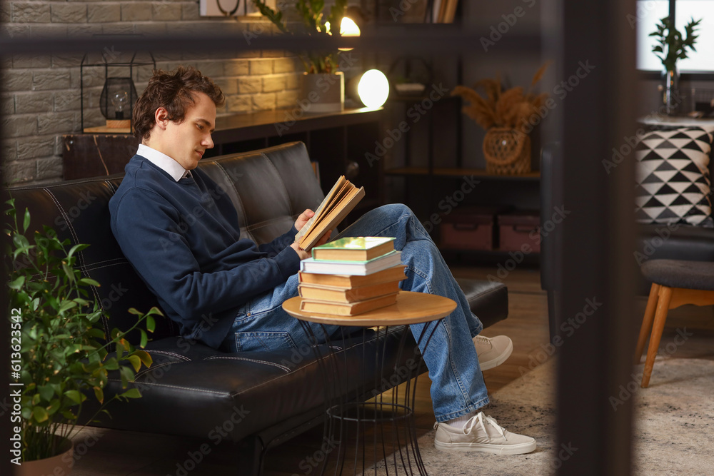 Young man reading book on sofa at home late in evening