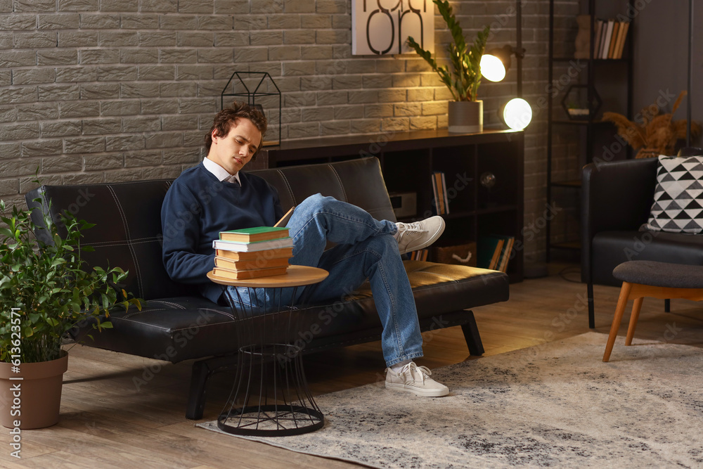 Young man reading book on sofa at home late in evening