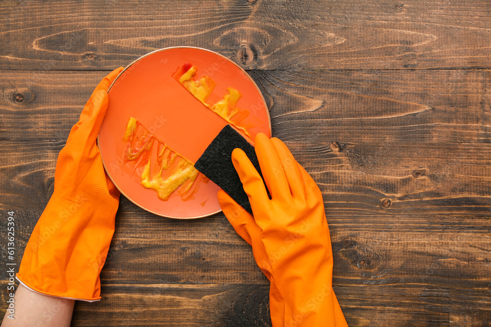 Female hands in rubber gloves washing dirty plate with sponge on wooden background