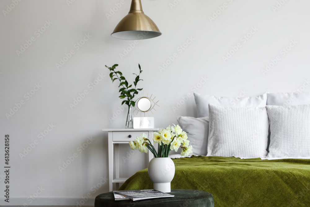 Interior of light bedroom with cozy bed and narcissus flowers on pouf