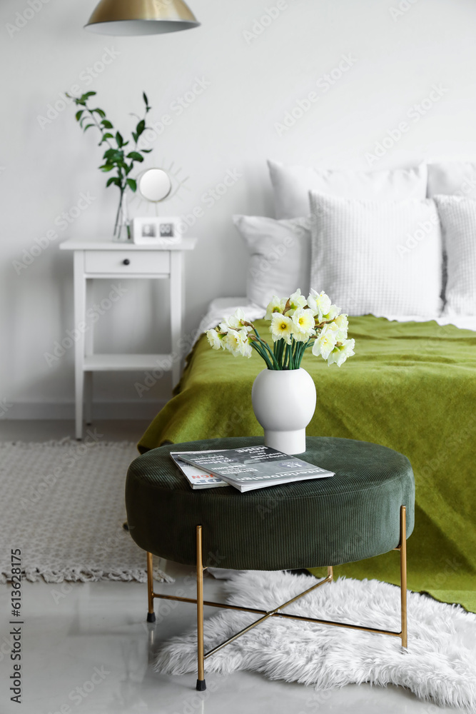 Interior of light bedroom with cozy bed and narcissus flowers on pouf