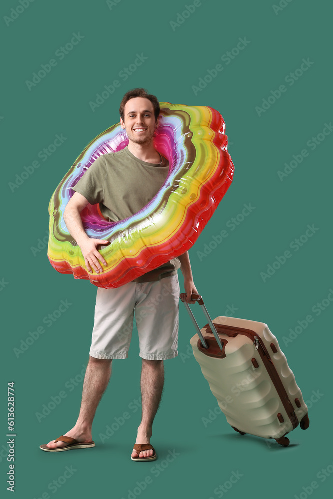 Young man with inflatable ring and suitcase on green background