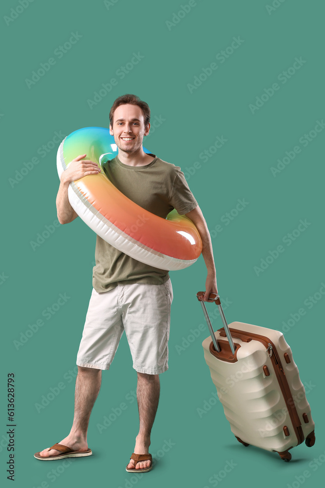 Young man with inflatable ring and suitcase on green background