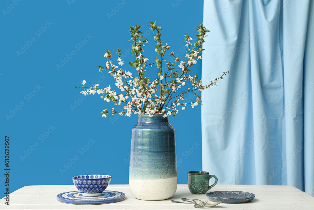 Vase with blooming tree branches and dishes on white table against blue background