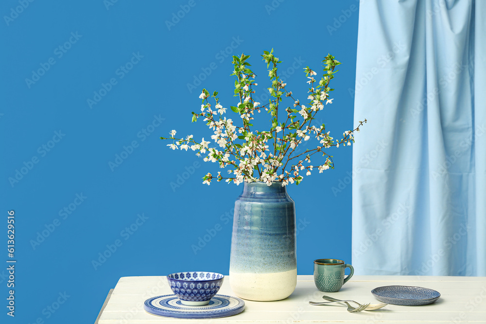 Vase with blooming tree branches and dishes on white table against blue background