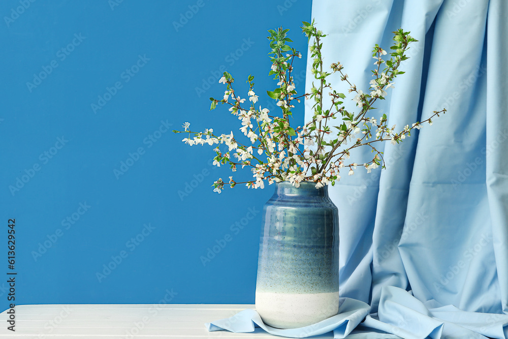 Vase with blooming tree branches and curtain on white table against blue background