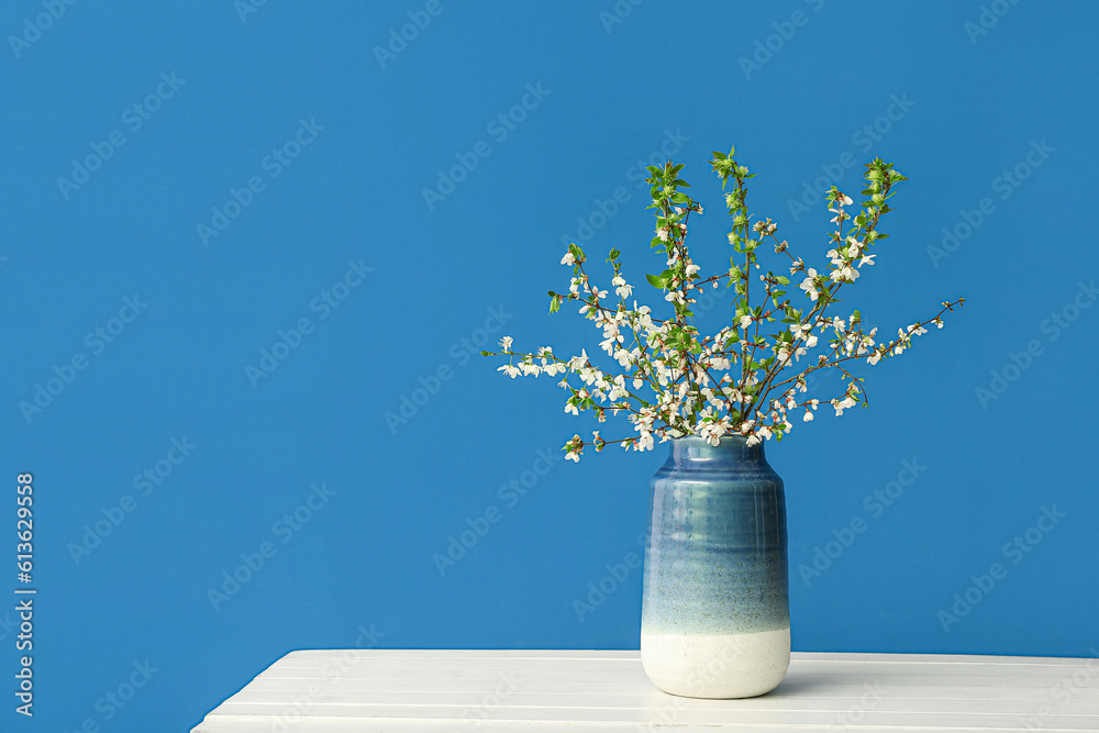 Vase with blooming tree branches on white table against blue background