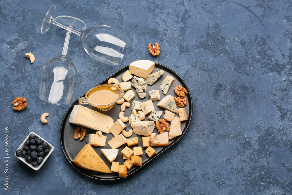 Tray with pieces of tasty cheese, honey and wine glasses on blue background