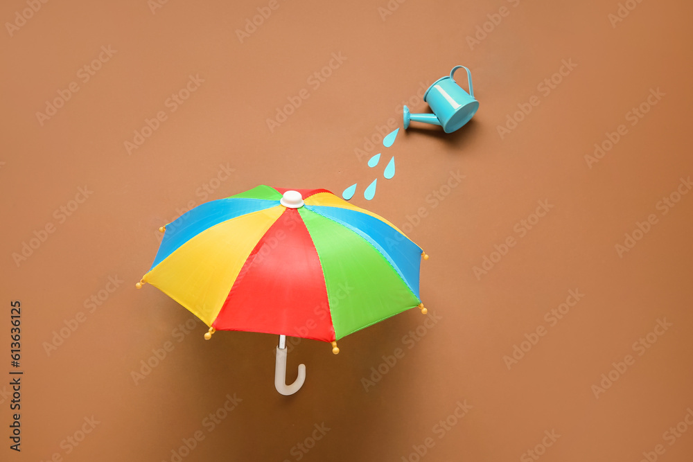 Creative composition with mini watering can and umbrella on brown background