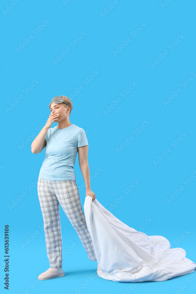 Young woman with soft blanket yawning on blue background