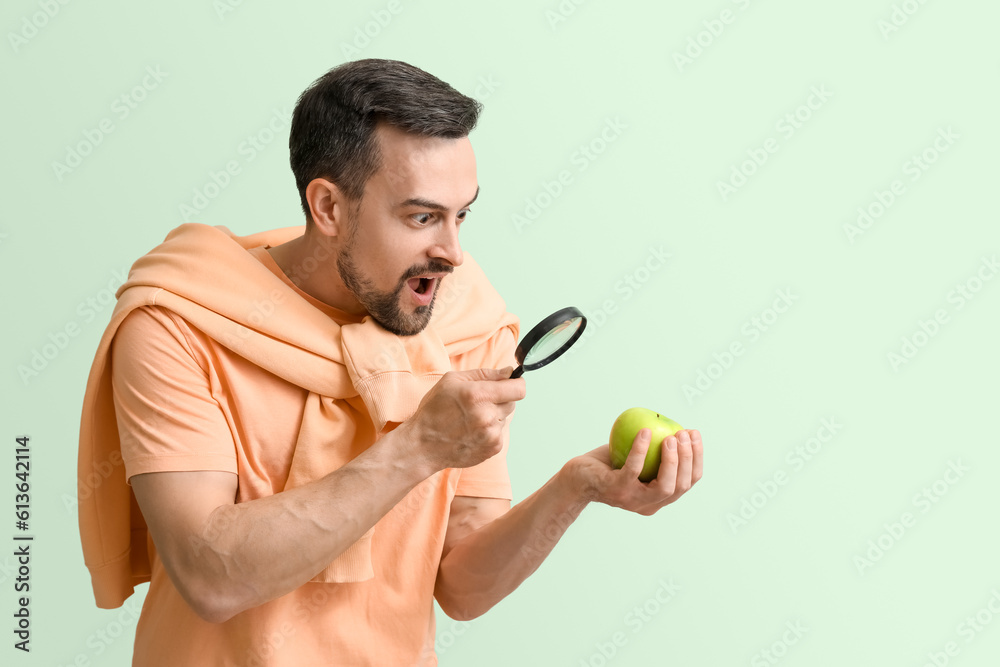 Surprised young man with magnifier and apple on green background