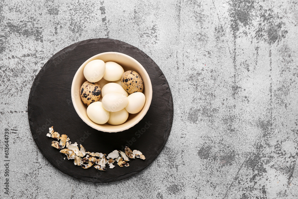 Board with bowl of boiled quail eggs and shells on grey background