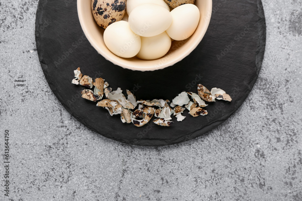 Board with bowl of boiled quail eggs and shells on grey background