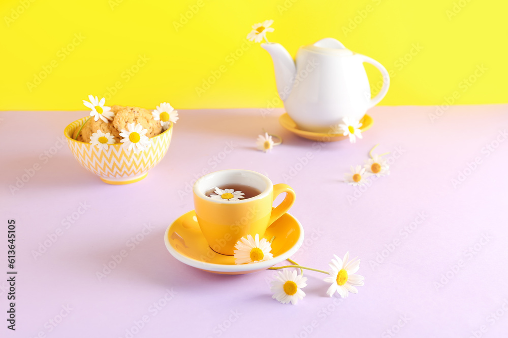 Teapot with cup of natural chamomile tea, cookies and flowers on pink table near yellow wall